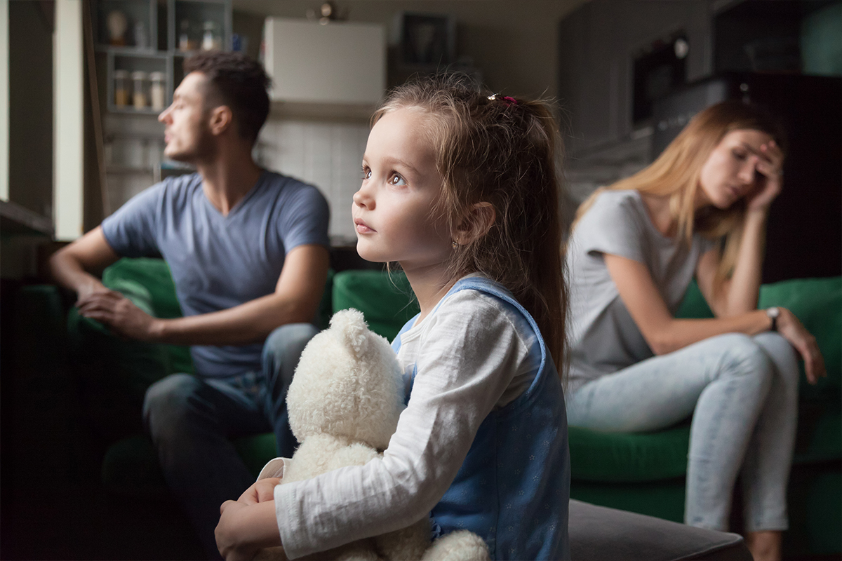 A young girl holding a teddy bear sits on a couch, looking pensively out a window as her parents sit apart, appearing distressed.