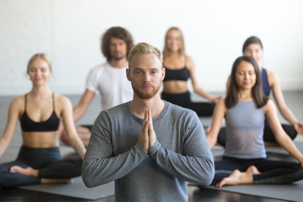 Group of young sporty people practicing Yin yoga lesson with instructor, sitting in Sukhasana exercise, Easy Seat pose, friends working out in club, focus on male student, making mudra gesture, studio