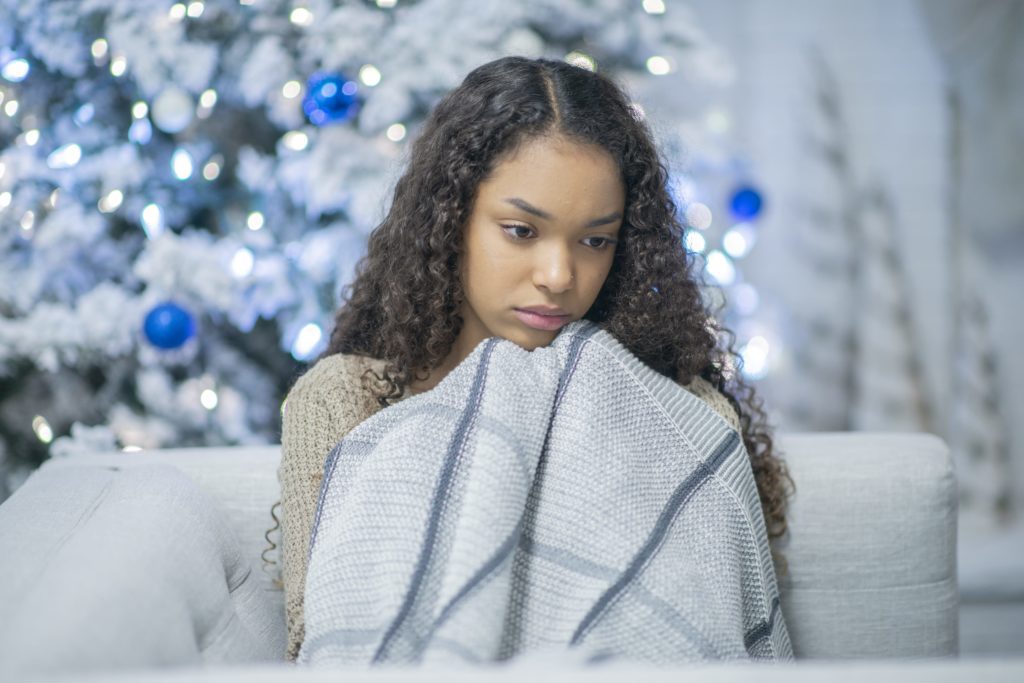 A lonely girl of african descent cuddles under a blanket by herself. She is sad and depressed to be by herself for the holidays.