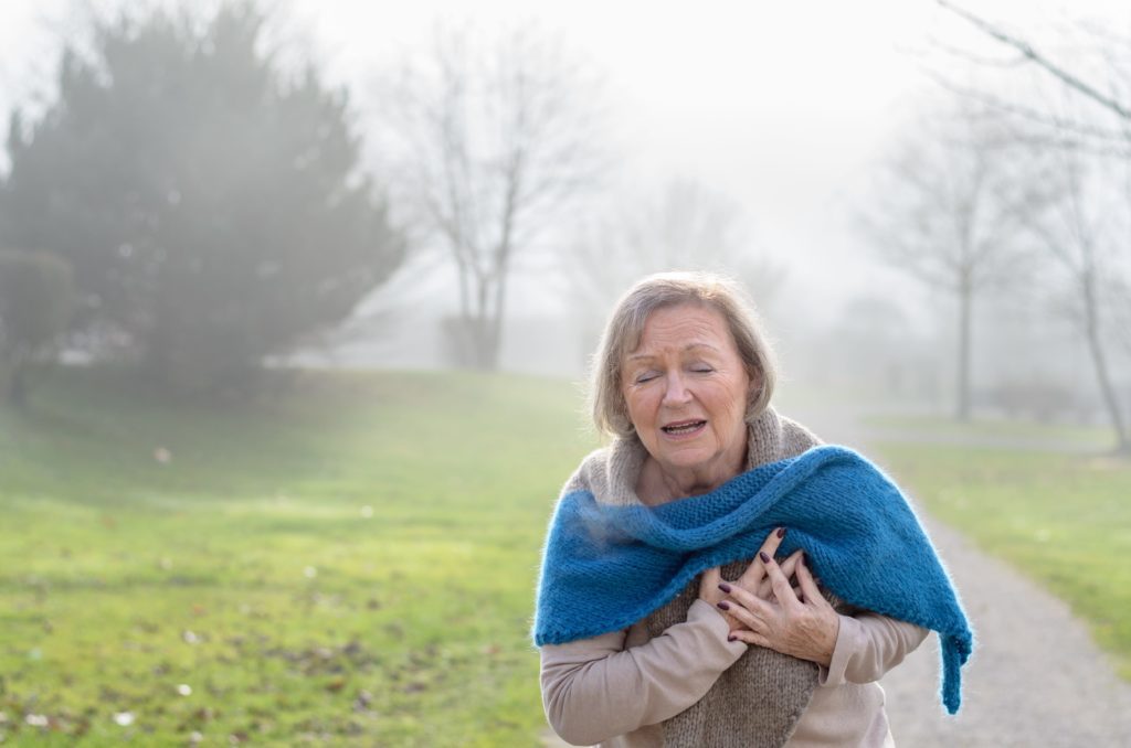 Senior lady clutching her chest in pain at the first signs of angina or a myocardial infarct or heart attack, upper body on a rural lane on a misty winter day