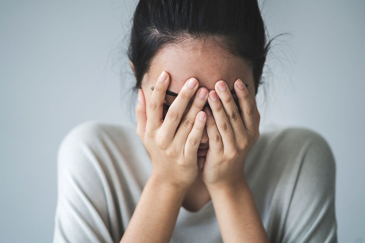 A person with long hair sits against a plain background, covering their face with hands, conveying distress or sadness.