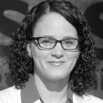 A woman with curly hair and glasses smiles at the camera, wearing a white shirt and a red collar against a blurred background.