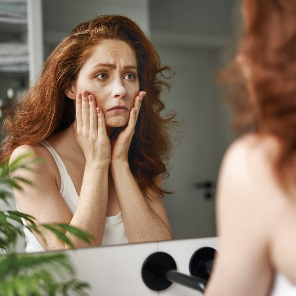 Woman with curly red hair gazes into a mirror, looking concerned while touching her face. Green plant in foreground.