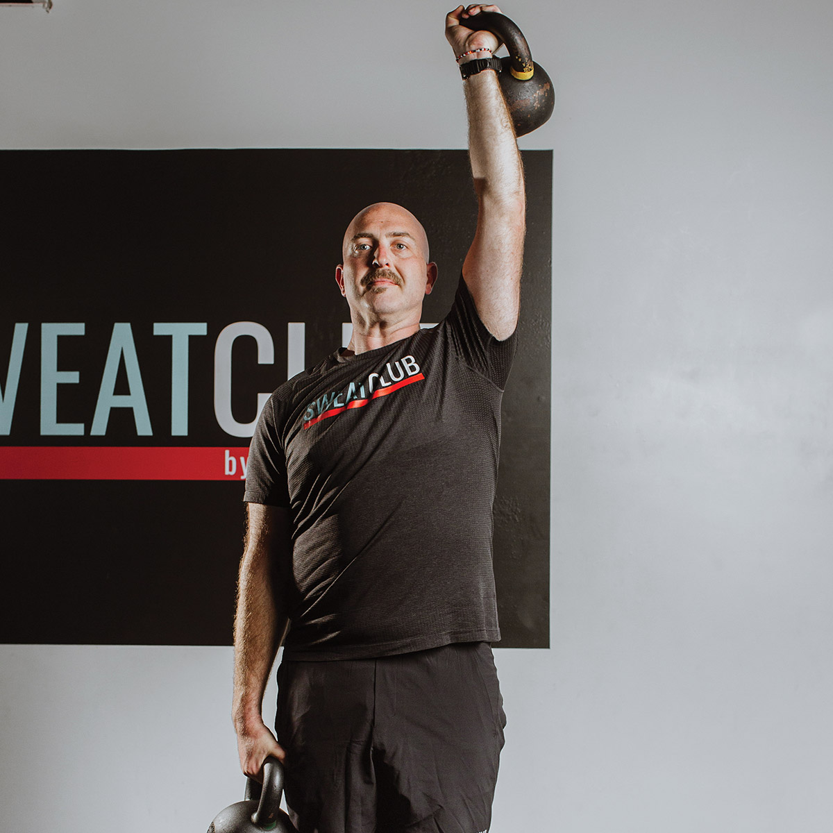 A man in a black t-shirt lifts a kettlebell overhead, with a gym logo on the wall behind him.