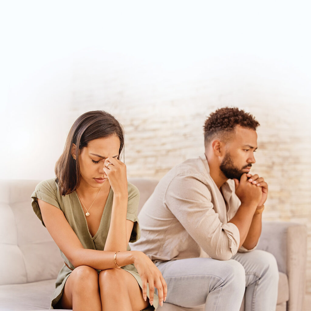 A couple sits apart on a couch, looking distressed and deep in thought, conveying tension and emotional distance.