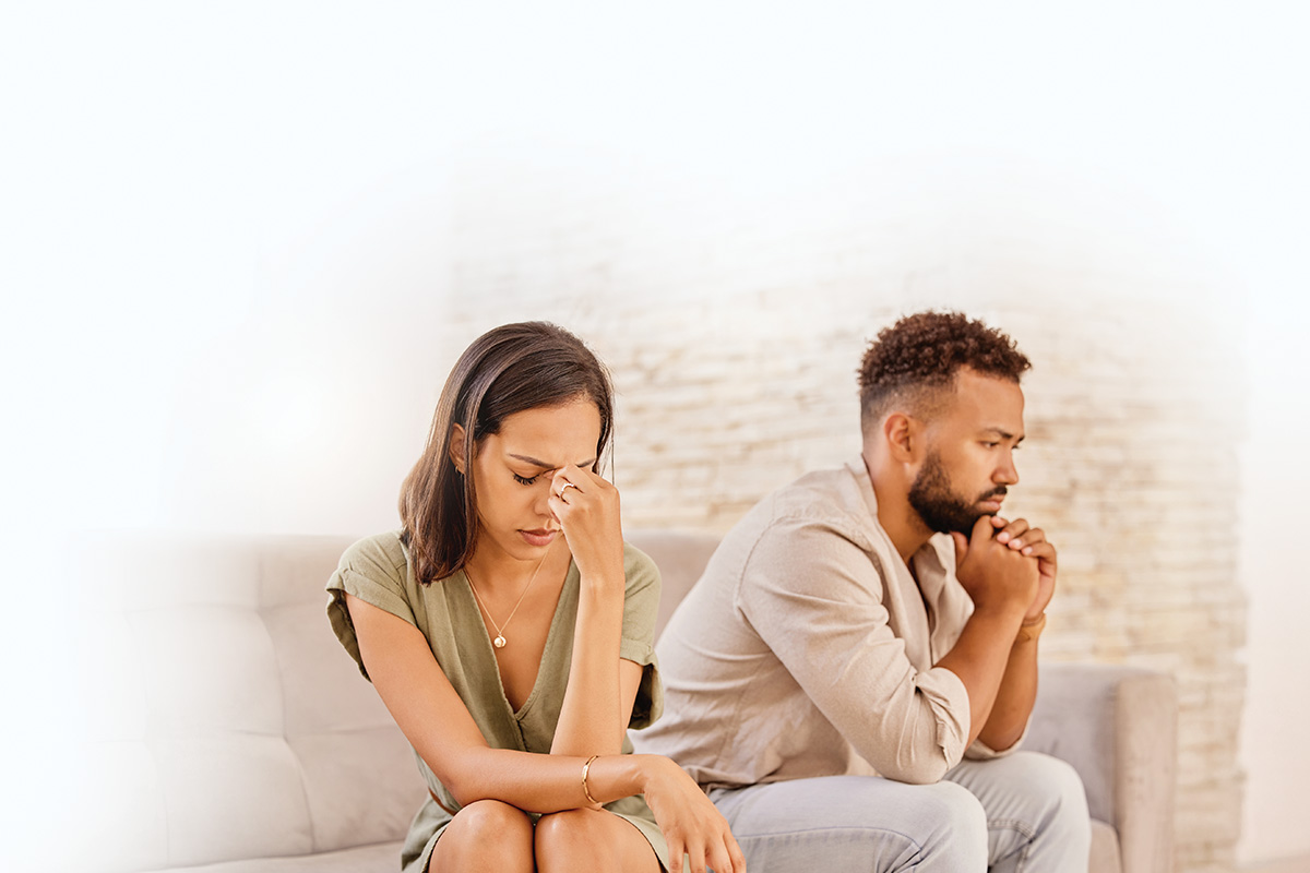 A couple sits apart on a couch, looking distressed and deep in thought, conveying tension and emotional distance.