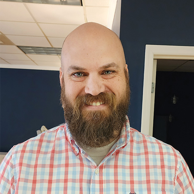 A smiling man with a bald head and a beard, wearing a checkered shirt, stands in an office setting with blue walls.