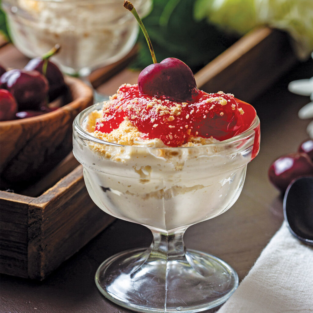 A glass dessert cup filled with whipped cream, cherry topping, and crushed nuts, garnished with a cherry on top.