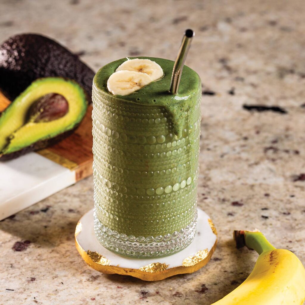 A green smoothie in a textured glass, topped with banana slices, beside an avocado and banana on a countertop.