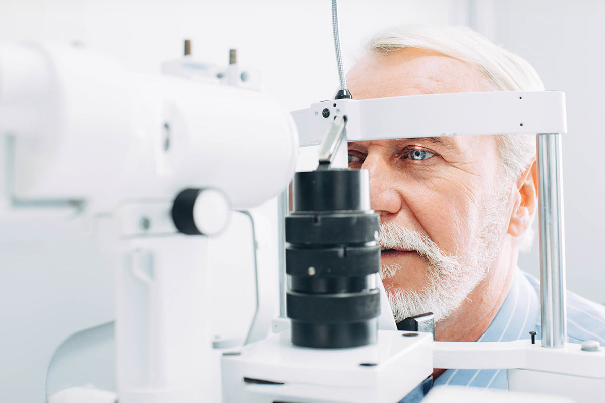 An elderly man with a beard undergoing an eye examination using specialized equipment in a bright clinical setting.