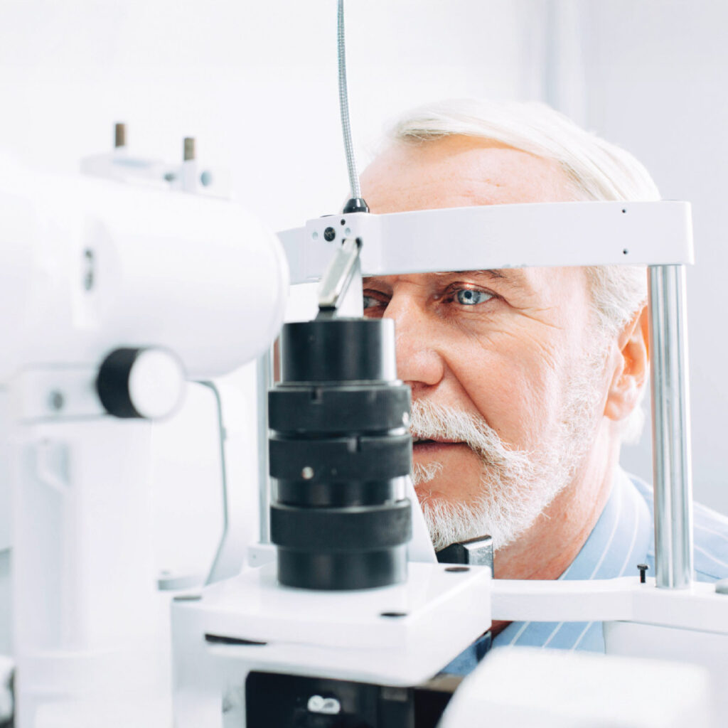 An elderly man with a beard undergoing an eye examination using specialized equipment in a bright clinical setting.