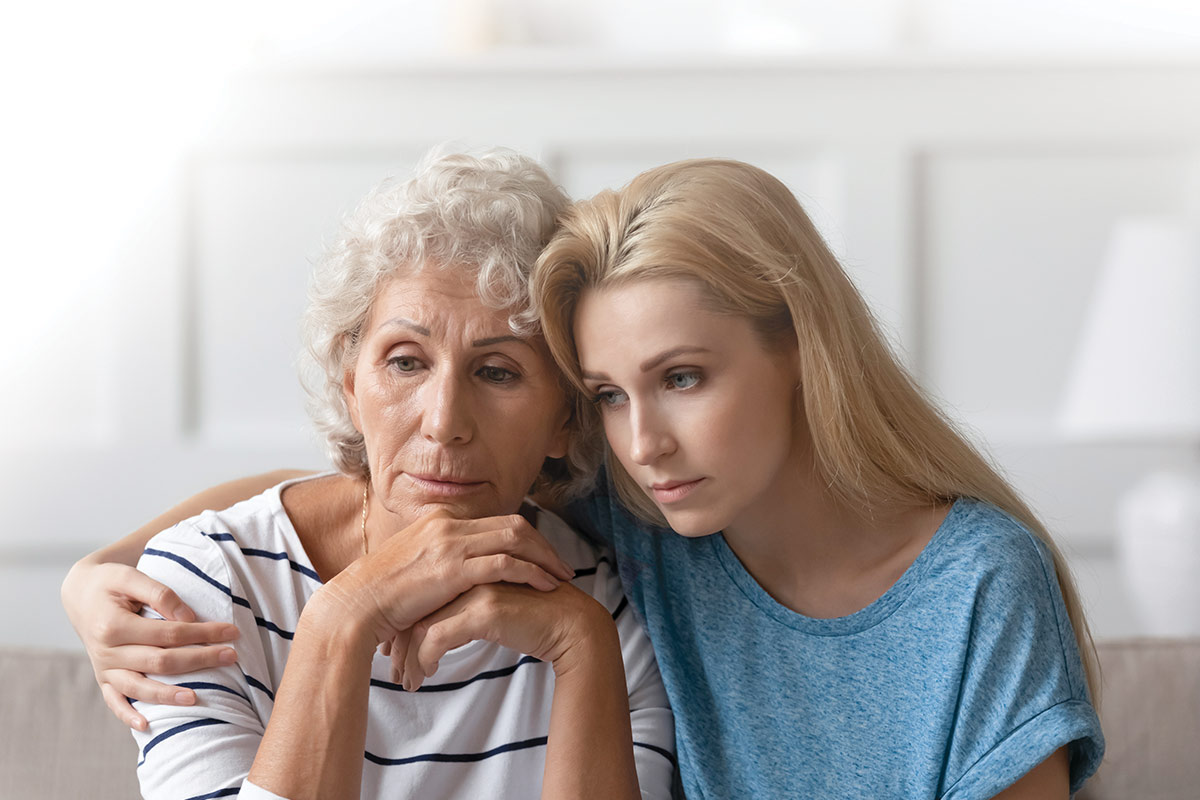 A young woman comforts an older woman, both looking concerned, sitting closely on a couch in a softly lit room.