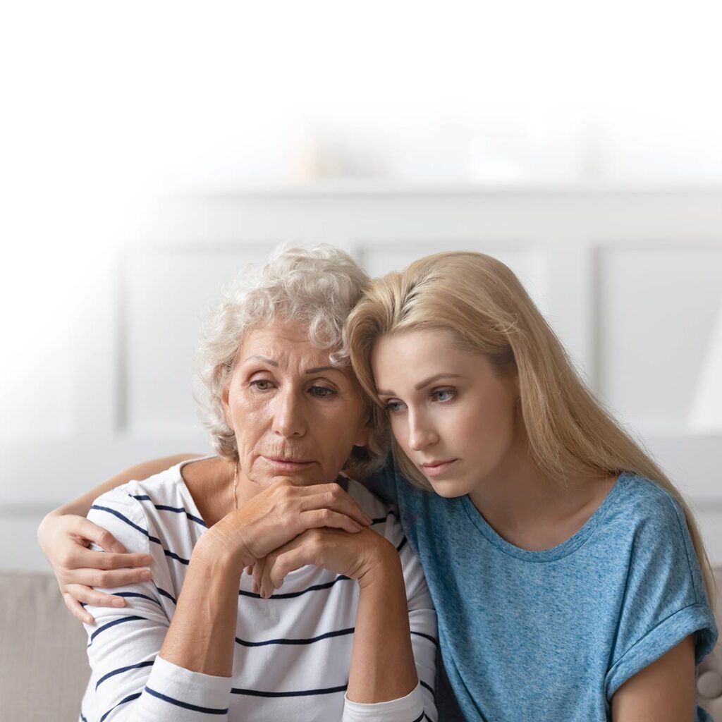 A young woman comforts an older woman, both looking concerned, sitting closely on a couch in a softly lit room.