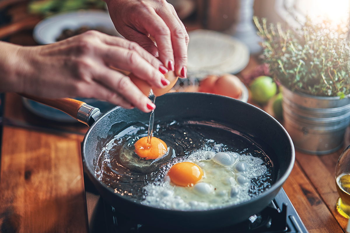 A hand cracking an egg into a frying pan on a wooden table, with herbs and ingredients in the background.