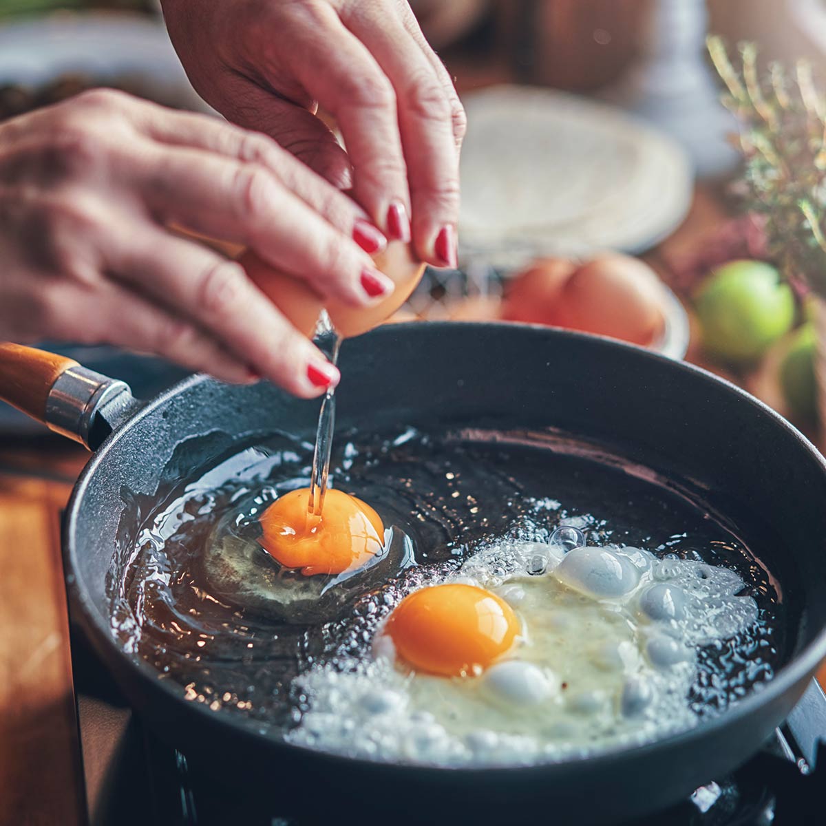 A hand cracking an egg into a frying pan on a wooden table, with herbs and ingredients in the background.