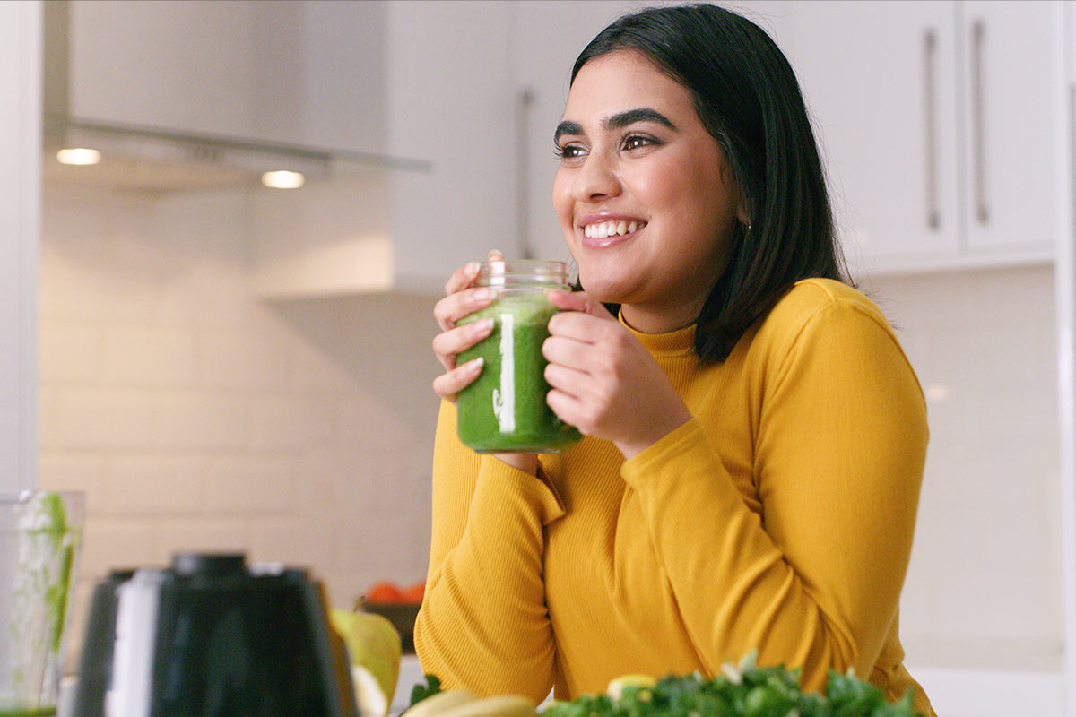 A woman in a yellow sweater smiles while holding a green smoothie in a bright kitchen filled with fresh ingredients.