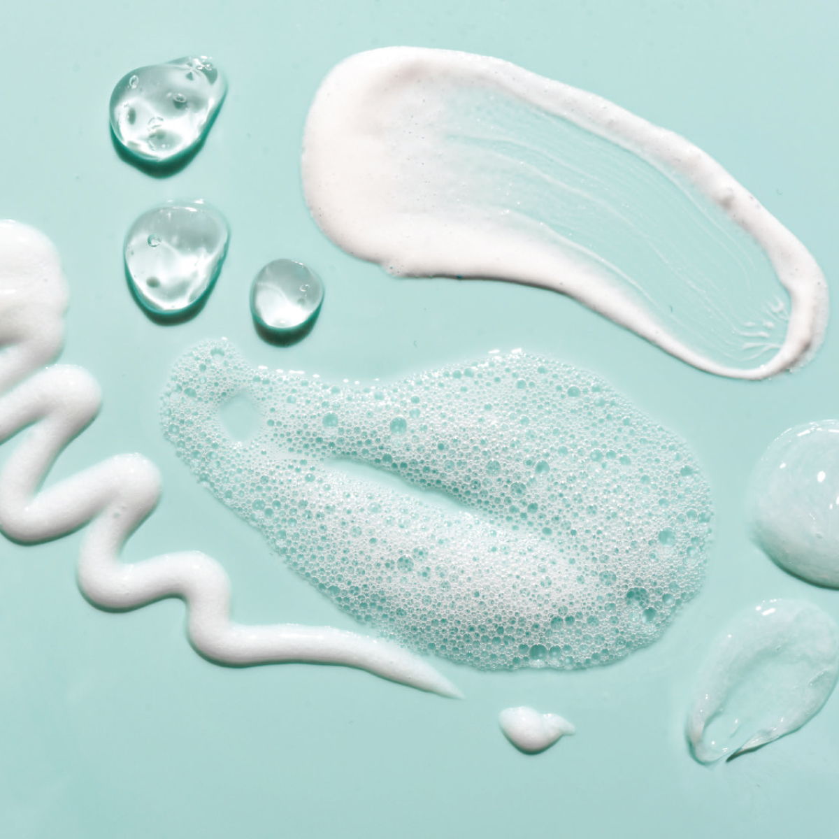 Various textures of blue and white skincare products on a soft turquoise background, including droplets and foam.