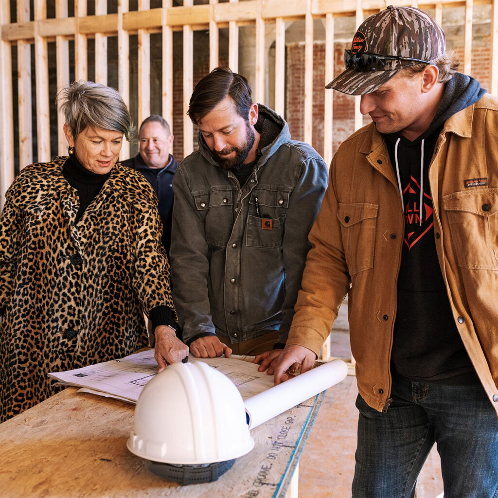Three people stand around blueprints with construction around them
